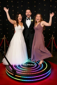 Bride, groom, and bridesmaid having fun on a Rumba 360 LED video booth at a wedding reception with red carpet and black backdrop with fairy lights.